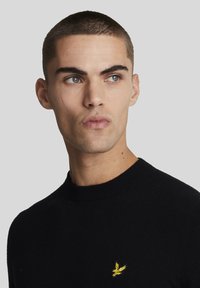 Fekete kötött pulóver kerek nyakkivágással, bal mellrészen egy kis sárga sas logóval. Sima textúra, testhezálló kialakítás.