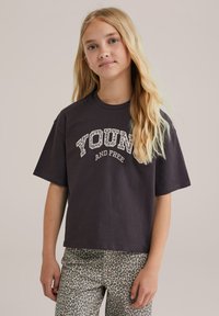 Tamsiai pilka trumpa rankovė marškinėliai su "JAUNAS IR LAISVAS" grafika tekstūrinėmis, baltomis raidėmis. Suderinta su pilkų leopardų rašto kelnėmis.