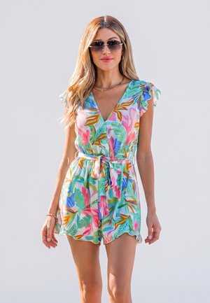 Femme aux longs cheveux blonds portant des lunettes de soleil et une combinaison courte fleurie colorée avec une ceinture, debout devant un fond clair.
