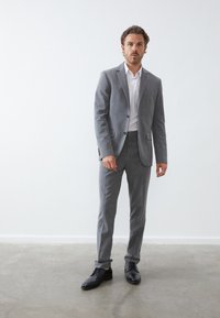 Grau maßgeschneiderter Anzug mit einem Ein-Knopf-Jacket, kombiniert mit einem weißen Dress-Shirt und schwarzen Schnürschuhen. Glattes Material und strukturierte Gestaltung.