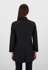 Cappotto nero su misura con una texture liscia, apertura centrale sul retro e polsini arrotolati, visto da dietro. Design semplice e strutturato.