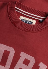 Close-up of a red Superdry sweatshirt showing ribbed neckline, white striped detail, and partial bold white brand lettering on fabric.