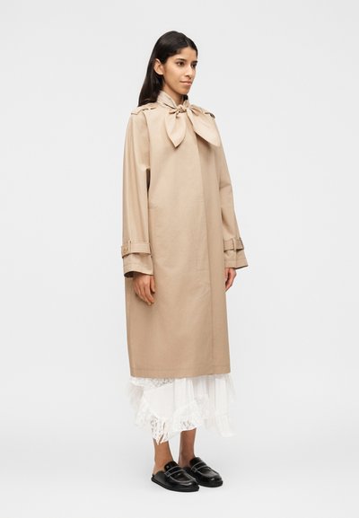 Γυναίκα όρθια,  φοράει ένα μακρύ μπεζ παλτό με μεγάλο φιόγκο στο λαιμό, μια λευκή δαντελένια φούστα και μαύρα παπούτσια χωρίς κορδόνια, μπροστά σε ένα απλό φόντο.