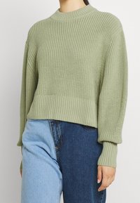 Pull vert clair en tricot avec un col rond côtelé et des poignets côtelés. Présente une coupe décontractée et un motif texturé. Porté avec un bas en denim mélangé.
