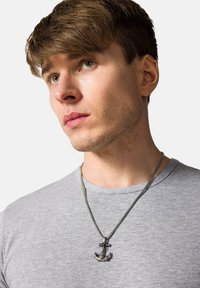 Srebrny wisiorek w kształcie kotwicy na łańcuszku, noszony na szarej koszulce. Wisiorek ma szczegółowe elementy wzornictwa morskiego.