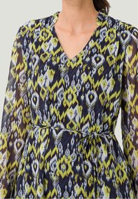 Femme portant une robe à manches longues et décolleté en V avec un motif géométrique jaune, marine et blanc, cintrée à la taille avec une ceinture en tissu assorti.