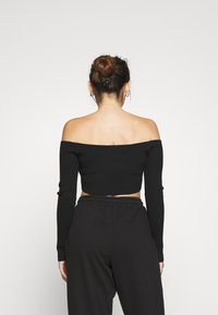Svart off-the-shoulder kort topp med långa ärmar, ribbad textur, i kombination med svarta byxor med elastisk midja, sedd bakifrån.