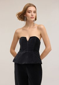 Kobieta z blond włosami do ramion, nosząca długie srebrne kolczyki z chwostami oraz czarny aksamitny top bez ramiączek z baskinką i dopasowane spodnie.