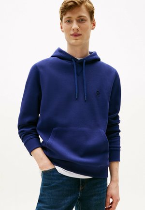 Jeune homme portant un sweat à capuche bleu avec une poche avant et un jean bleu foncé, debout avec une main dans la poche.