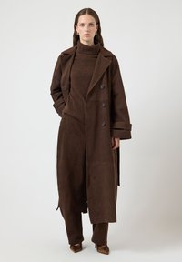Długi brązowy zamszowy płaszcz z podwójnym zapięciem, dużymi klapami i kieszeniami, zestawiony z dopasowanym brązowym dzianinowym golfem i spodniami.