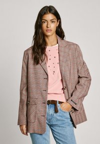 Blazer a cuadros marrón con un corte relajado, que presenta detalles en rosa claro, dos botones y un bolsillo. Combinado con una blusa rosa clara y jeans azules.