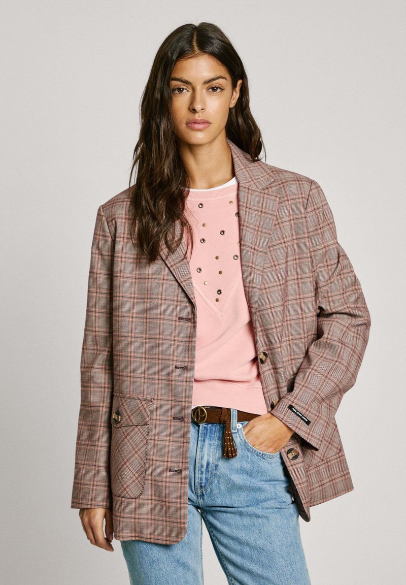 Blazer a cuadros marrón con un corte relajado, que presenta detalles en rosa claro, dos botones y un bolsillo. Combinado con una blusa rosa clara y jeans azules.