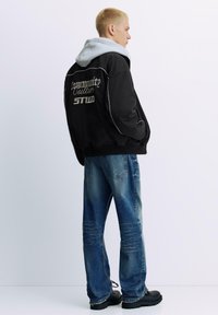 Zwarte bomberjack met witte details en tekst op de achterkant, gedragen over een grijze hoodie; gecombineerd met versleten blauwe jeans en zwarte laarzen.