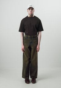 Ruda trumpa rankovėliams marškinėliai, laisvi alyvuogių žalumo džinsai ir rudi batai. Balta skrybėlė su juodu tekstu. Modelis stovi prieš paprastą fono.