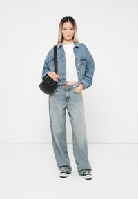 Veste en denim bleu clair avec boutons, t-shirt court blanc, jeans larges bleu clair, sac bandoulière noir et baskets noires.