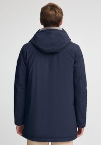 Giubbotto imbottito blu navy con cappuccio, dotato di chiusura a zip e tasche laterali. Texture liscia con una finitura leggermente lucida.