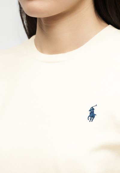 Κοντινό σε μια γυναίκα που φοράει ένα κρεμ πουλόβερ με ένα μικρό μπλε κεντημένο λογότυπο παίκτη πόλο στο στήθος.