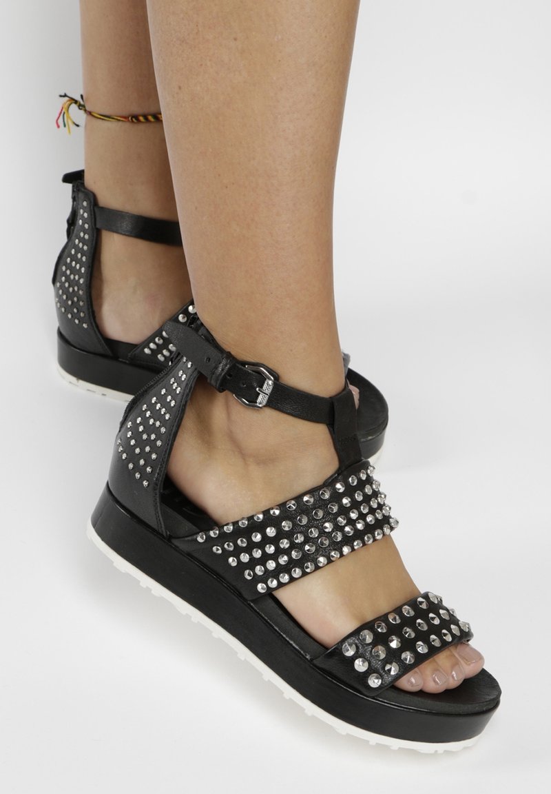 Sandales plateformes noires avec des clous argentés, bride à la cheville avec boucle, portées pieds nus avec un bracelet de cheville multicolore sur une jambe.