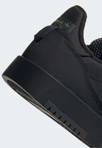 Sneaker nera con tomaia in rete testurizzata, pannelli sintetici lisci e suola in gomma rigata. Presenta un logo sulla linguetta e dettagli sul tallone.