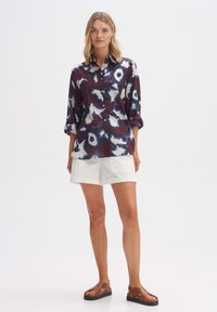 Hemd mit Knöpfen in dunkelrot und blauem abstraktem Muster, langärmlig hochgekrempelt, kombiniert mit weißen Shorts und braunen Sandalen.