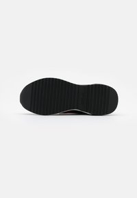 Semelle d'une chaussure noire avec des rainures horizontales et des zones texturées au talon et aux orteils sur un fond clair uni.