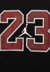 Czarna tkanina ozdobiona jest odważnymi czerwonymi cyframi "23" o teksturze przypominającej skórę gada. Biały logo Jumpman znajduje się na środku poniżej cyfr, z białą nicią jako obramowaniem.