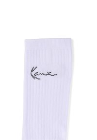 Weißer gerippter Socken mit einem schwarzen, kursiven Logo, das nahe am oberen Rand auf einem schlichten weißen Hintergrund gestickt ist.