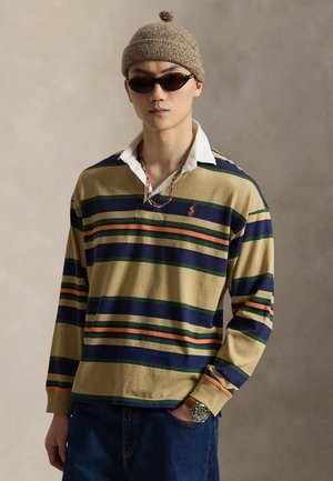 Jeune homme portant un maillot de rugby rayé, un bonnet en tricot marron avec pompon, des lunettes de soleil noires, un collier coloré en perles, un jean bleu et une montre-bracelet.