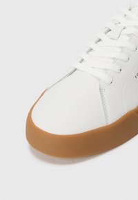 Białe skórzane sneakersy z białymi sznurowadłami i grubą brązową gumową podeszwą, z widocznymi detalami szycia i teksturowaną powierzchnią podeszwy.