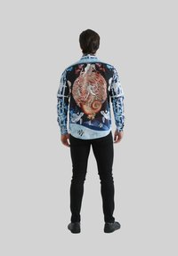 Homme de dos, portant une chemise à manches longues ornée de motifs colorés de la faune marine et de coquillages, associée à un pantalon noir et des chaussures.