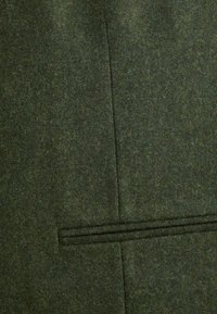 Tissu en laine vert foncé avec une texture lisse et de subtils flocons, présentant un design sur mesure et un détail de poche horizontale.