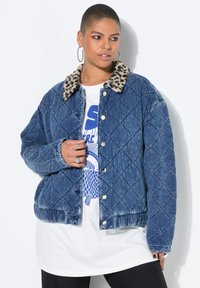 Pikowana kurtka dżinsowa z kołnierzem w panterkę, ozdobiona srebrnymi zatrzaskami i luźnym krojem. Noszona na białej koszulce z nadrukiem.