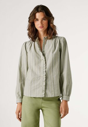 Jeune femme portant un chemisier à rayures vertes et blanches avec des détails à volants, associé à un pantalon vert clair, debout sur un fond blanc.