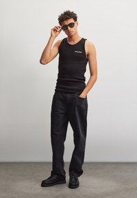 Jonge man in een zwart hemd, losse zwarte jeans en zwarte schoenen poseert binnen, draagt een zonnebril en past deze aan met één hand, de andere hand in zijn zak.
