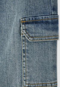 Jasnoniebieska dżinsowa tkanina z łatą kieszeniową w pomarańczowym przeszyciu i teksturowanym wykończeniem. Kieszeń ma lekko wypukły projekt.