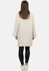 Knie-lange crème breigoed cardigan met lange mouwen en ribgebreide manchetten, gedragen over zwarte leggings en witte sneakers. Effen textuur, geen patronen.