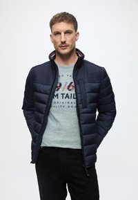 Sötétkék steppelt dzseki magas gallérral és vízszintes steppeléssel. Világosszürke pulóver alatt, nyomtatott grafikákkal. Fekete nadrág.