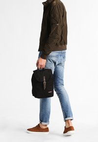 Black canvas backpack with a fold-over flap and buckle closure. Features a small logo on the front, carried by a person in casual attire.