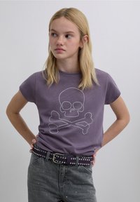 Tricou mov din bumbac cu un design de craniu și oase încrucișate realizat din nituri argintii. Modelul poartă o centură neagră cu nituri și blugi gri.
