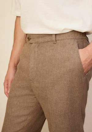 Close-up of person wearing textured brown trousers with a buttoned waistband and hand in right pocket, paired with a tucked white shirt.