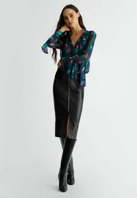 Blusa verde trasparente con motivo floreale rosa e maniche svasate, abbinata a una gonna a tubino nera in pelle aderente con zip frontale e spacco.