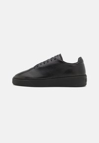 Black leather sneaker with a smooth finish, rounded toe, flat lace-up design, and a thick black rubber sole. Minimalist style and clean lines.