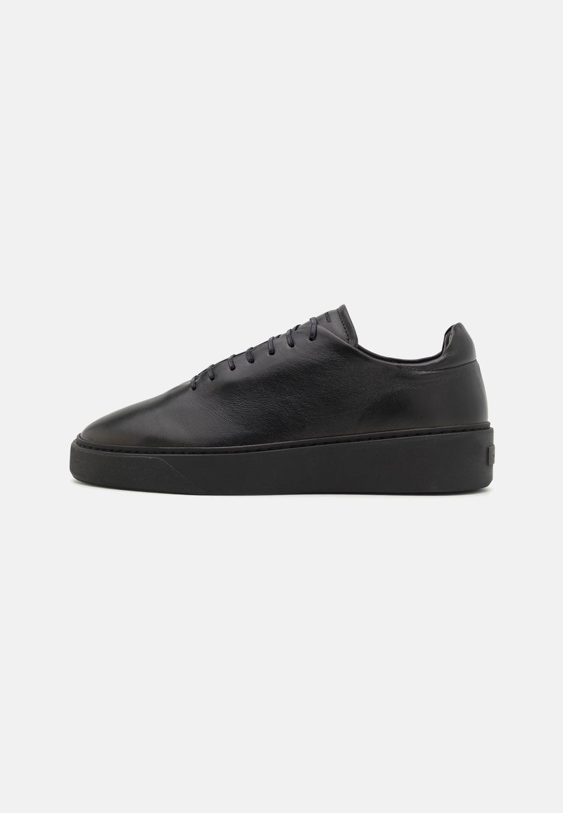Black leather sneaker with a smooth finish, rounded toe, flat lace-up design, and a thick black rubber sole. Minimalist style and clean lines.