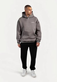 Felpa grigia con cappuccio regolabile e tasca frontale, abbinata a pantaloni neri e sneakers bianche. Tessuto liscio, design casual.