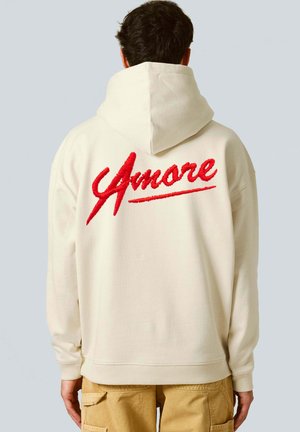 Cream hoodie with a textured red "Amore" graphic on the back. Features a spacious hood and ribbed cuffs, paired with tan cargo pants.
