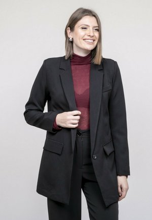 Jeune femme souriante, portant un blazer noir, un col roulé bordeaux et un pantalon noir, debout devant un fond clair uni.