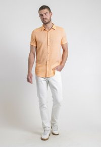 Chemise à manches courtes à boutons en tissu texturé orange clair, associée à un jean blanc et des baskets blanches avec des semelles beige.