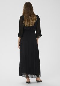 Čierne plisované maxi šaty s priesvitnými dlhými rukávmi a elastickým pásom, ktoré majú voľný strih a priame ukončenie.