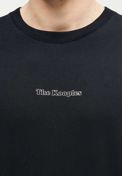Μαύρο μπλουζάκι με στρογγυλή λαιμόκοψη και λευκό κείμενο με περίγραμμα "The Kooples" στο κέντρο του στήθους, που φοριέται από άτομο ορατό από το λαιμό έως το άνω μέρος του κορμού.
