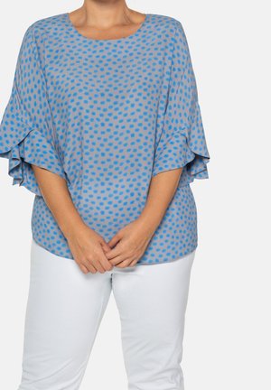 Vrouw draagt een lichtblauwe blouse met blauwe stippen en gelaagde mouwen, gecombineerd met een witte broek, staand tegen een effen achtergrond.